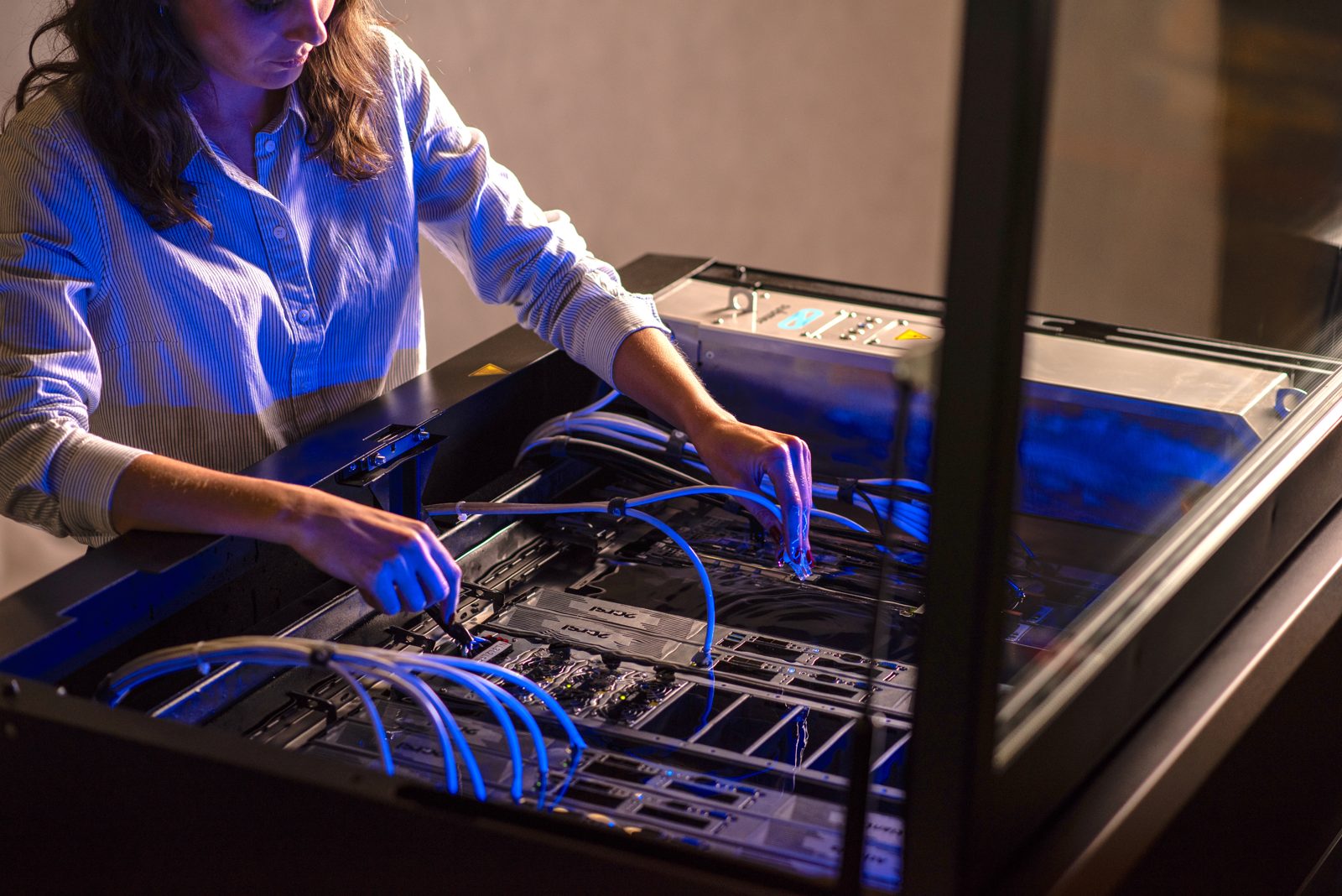 A technician lifting a server out of a Submer SmartPodX immersion tank, with blue cabling visible beneath the fluid